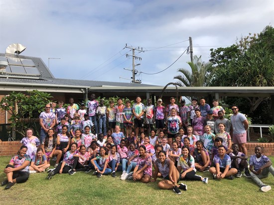 Large group of students and staff gathered outdoors in front of a school building, wearing colourful clothing and sitting and standing on grass lawn